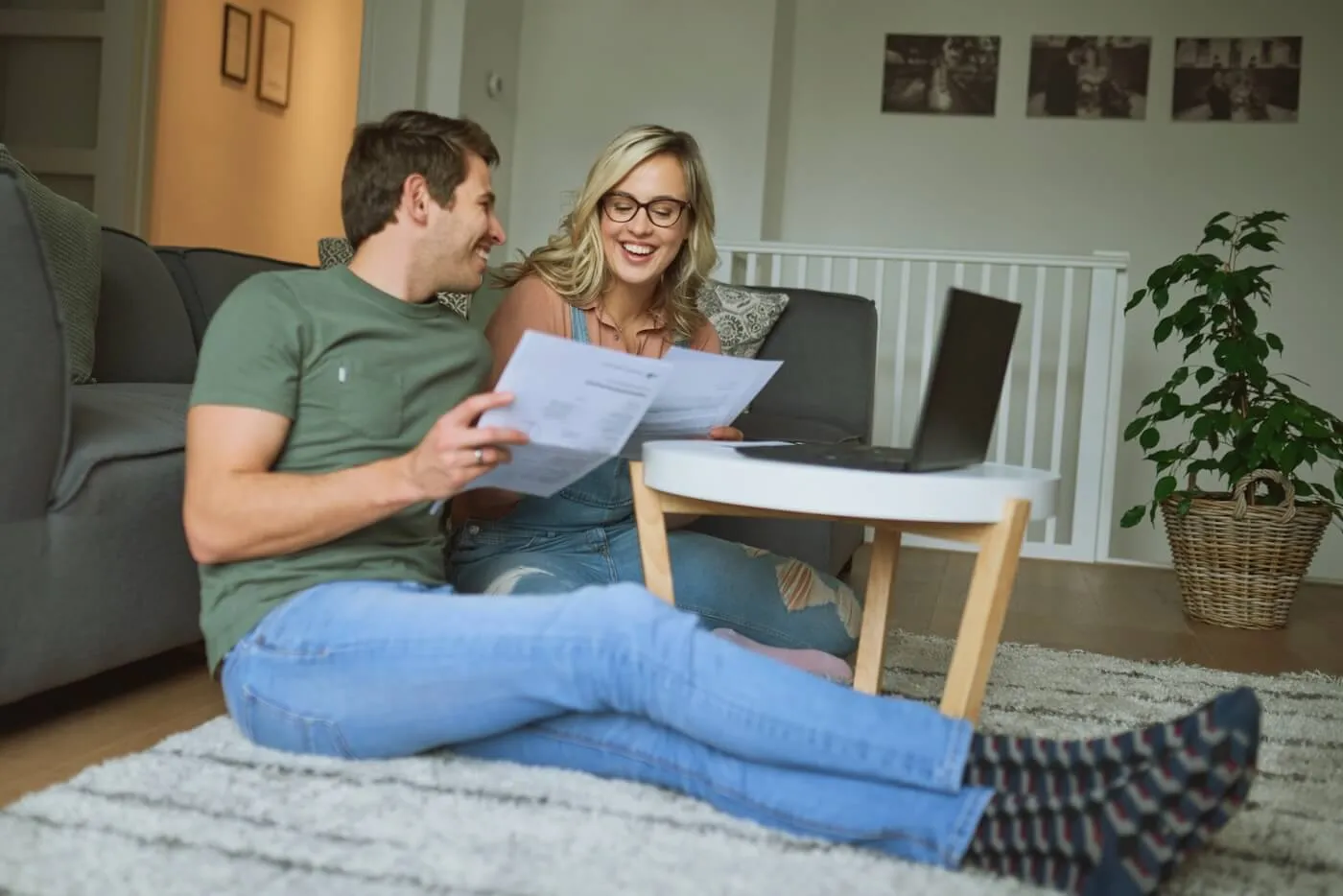 Happy young couple sitting on the rug in the living room, reviewing the bills and using the laptop