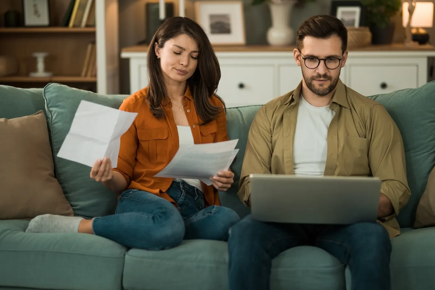 A man and a woman sitting on a couch holding papers and using a laptop to create a savings plan.