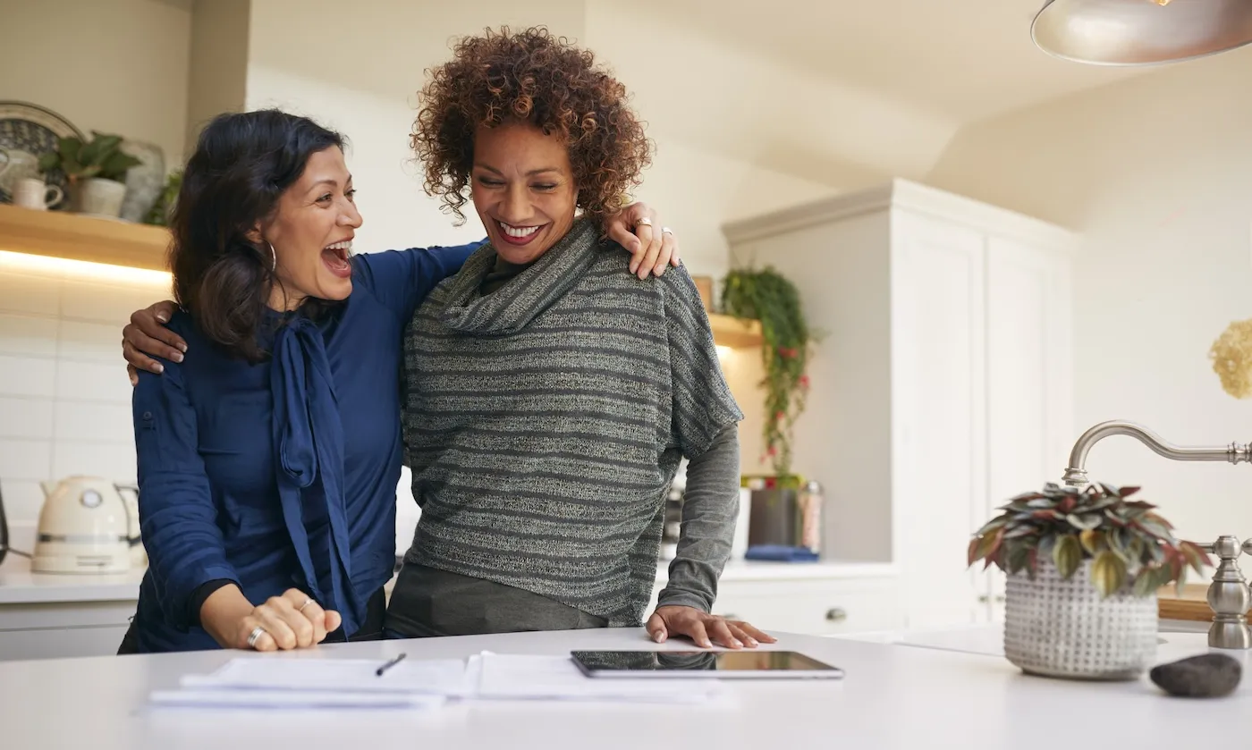 A smiling woman embracing her wife while reviewing their finances in the kitchen at home.