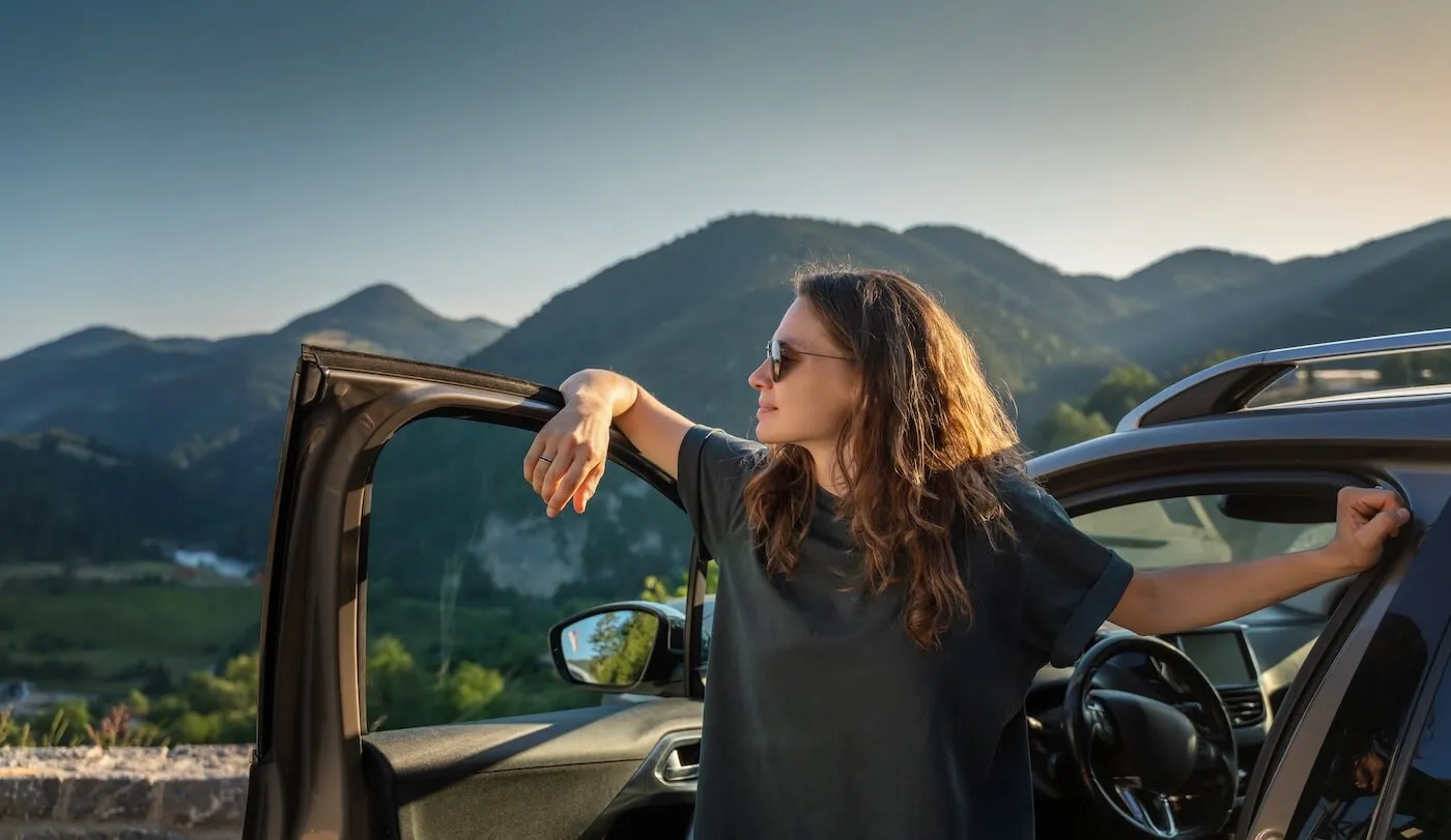 Young woman enjoys the mountain view while standing next to her car