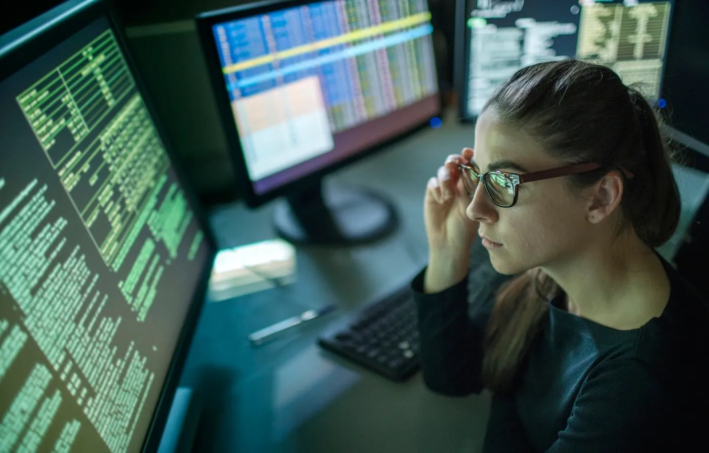 Focused woman in glasses is looking at the code on her monitors while sitting in the dark office