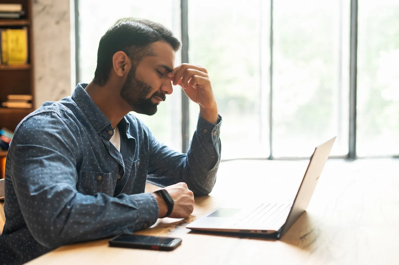 An exhausted man dealing with bankruptcy sits at his laptop and pinches his nose bridge with eyes closed.