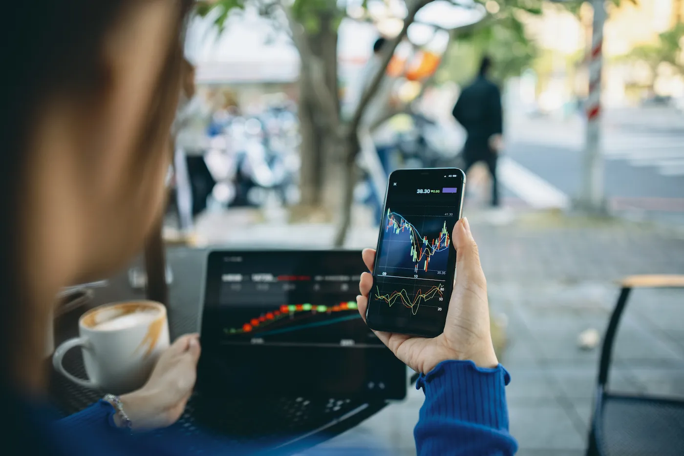 Person sitting outside at a cafe with a laptop and a mobile phone looking at stock performances.