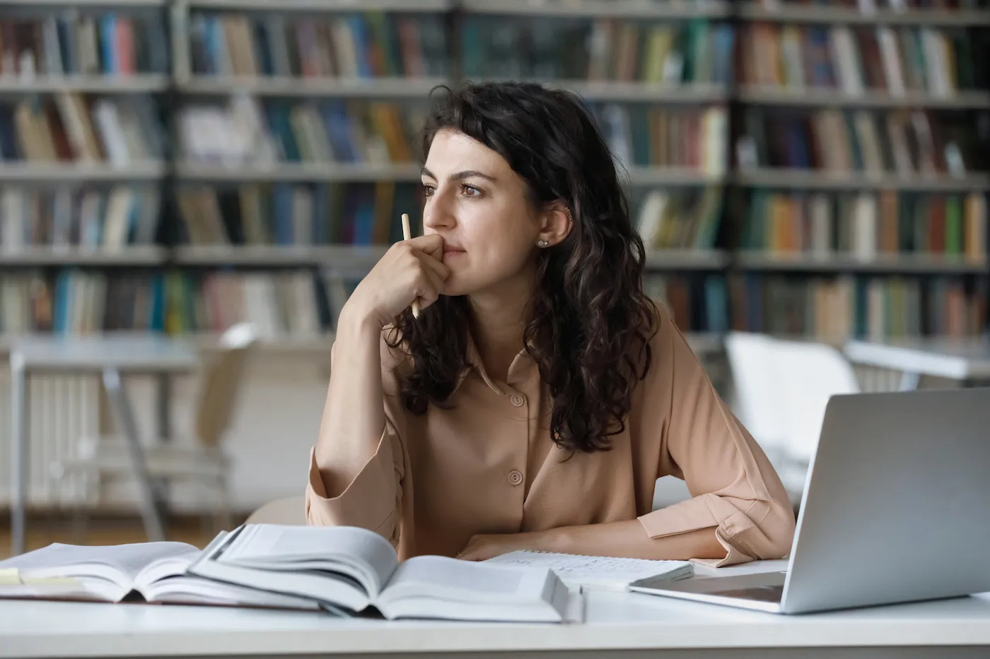 Young woman sit at table with textbooks and laptop staring aside, studying alone in library.