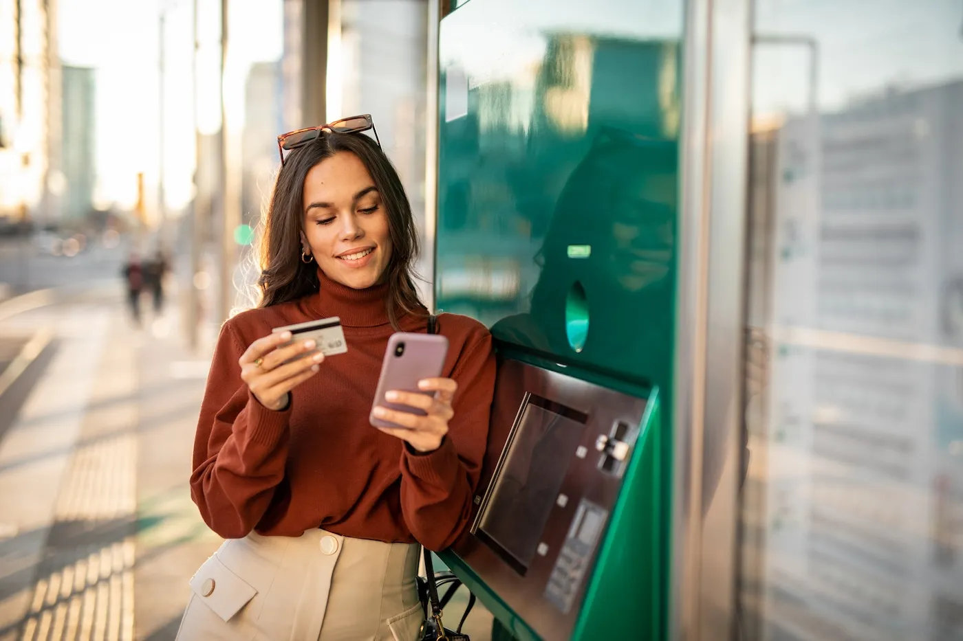 A young woman stopped by an ATM holding her credit card and phone.