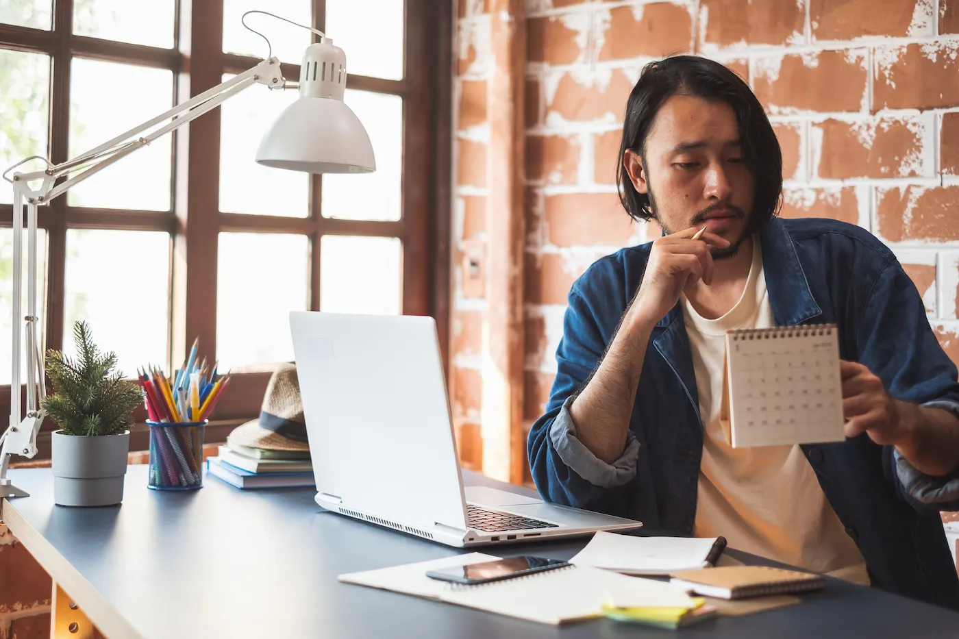 A man holding and looking at a paper calendar while sitting at his desk with a laptop.