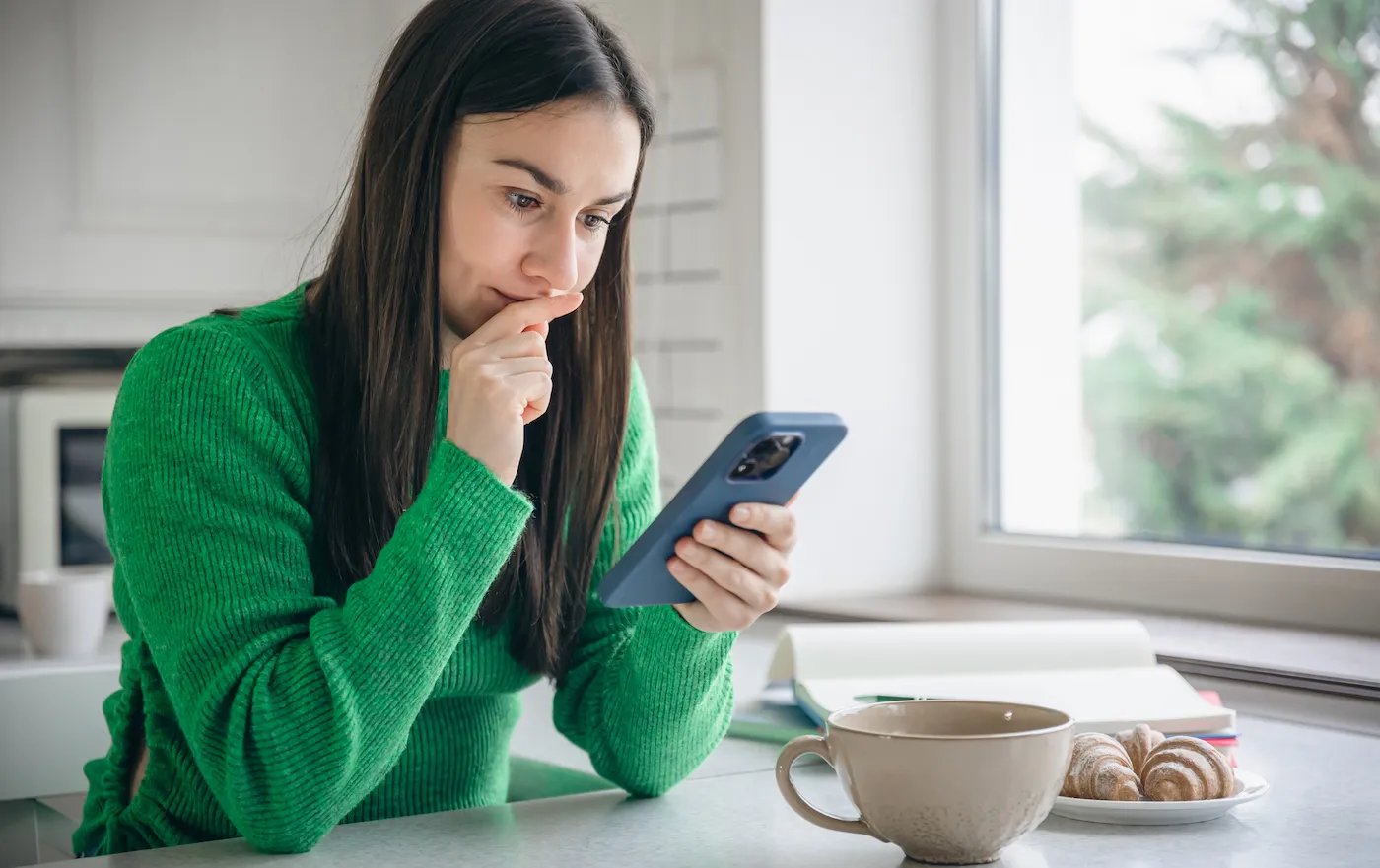 A woman in the morning in the kitchen checking her savings account with a smartphone and enjoying a cup of coffee.