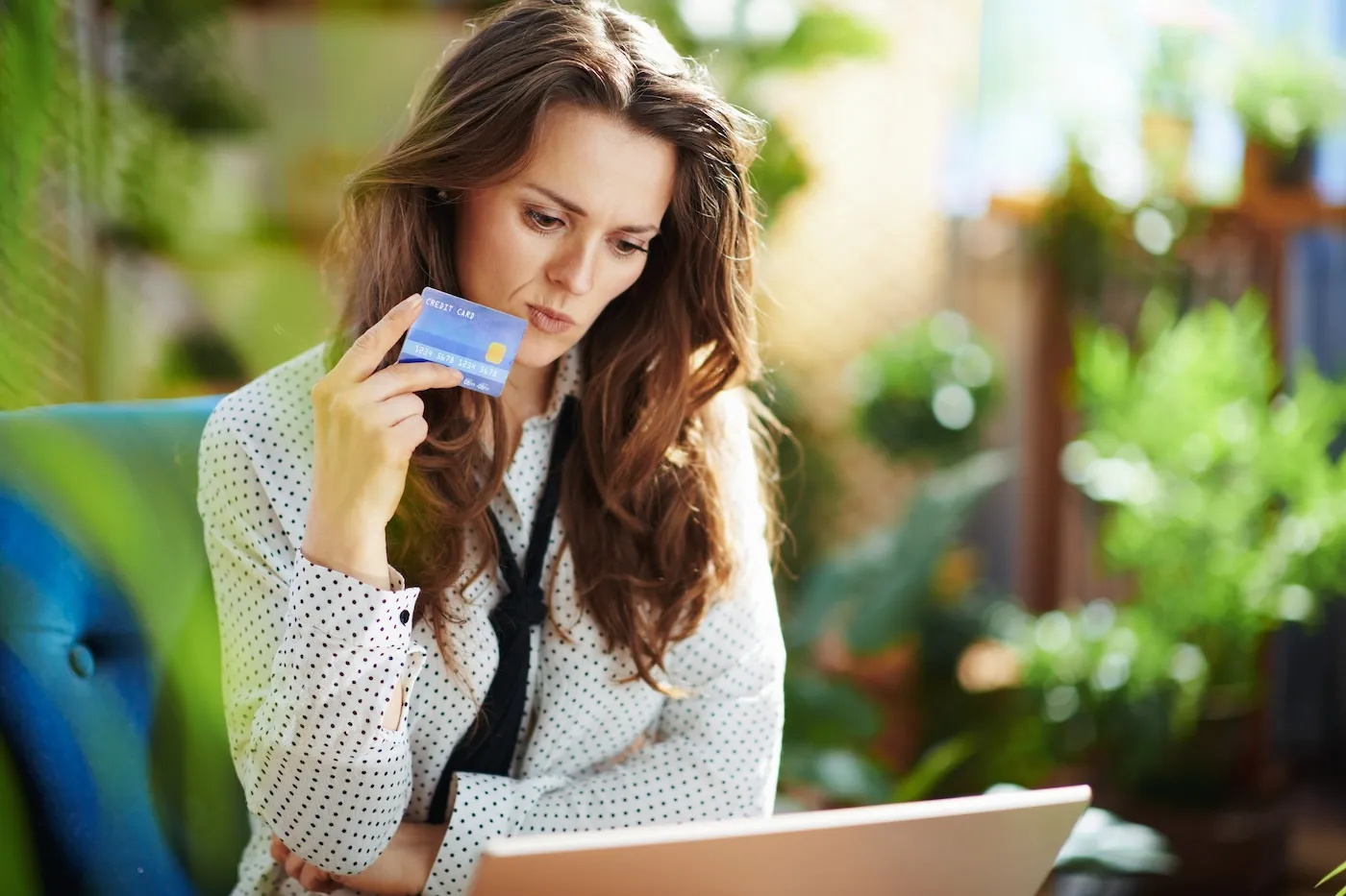 A woman with laptop and credit card sitting in a blue armchair in the green living room in sunny day.