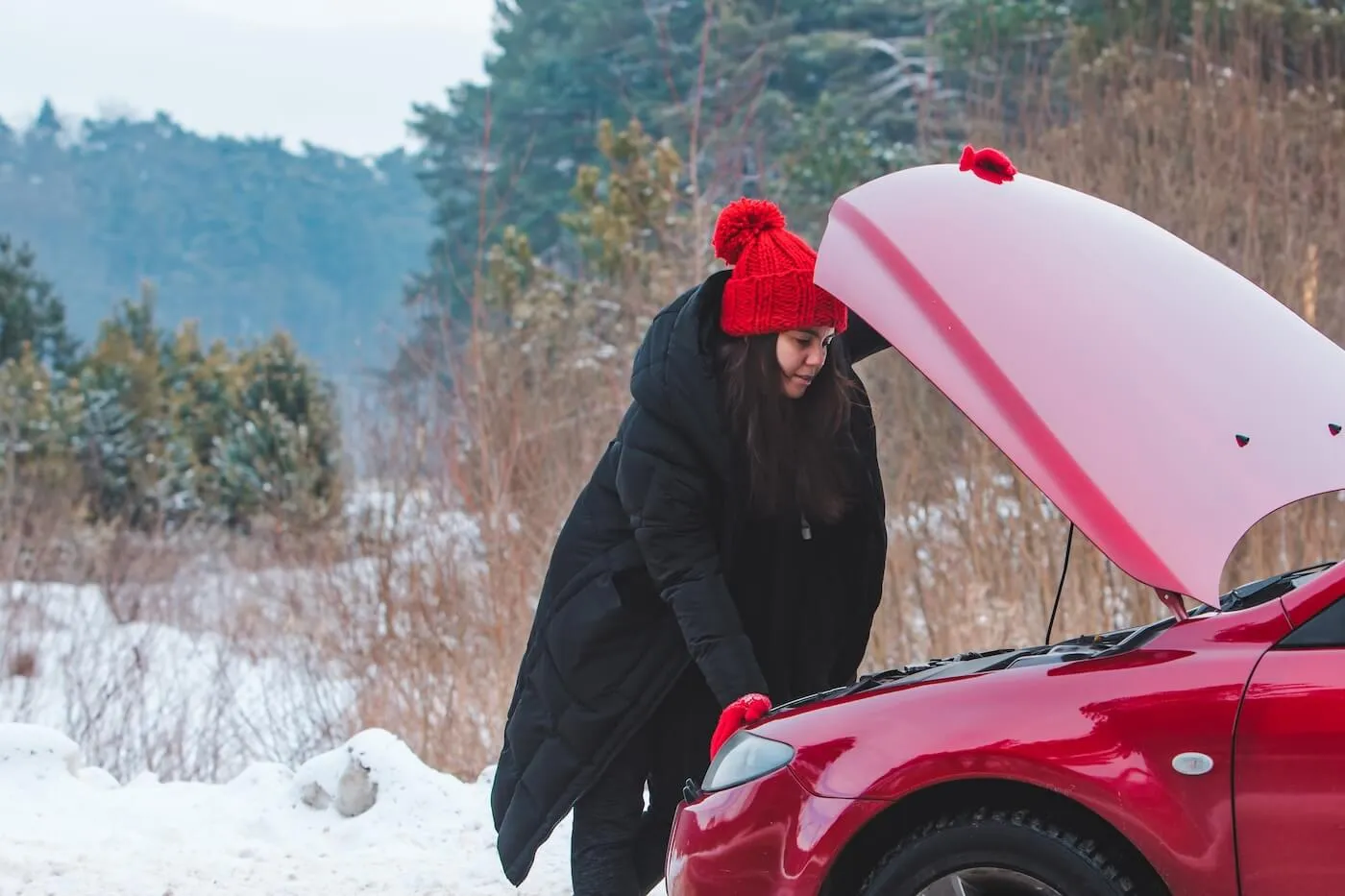 Young woman in winter outfit is looking under the car hood on a snowy street