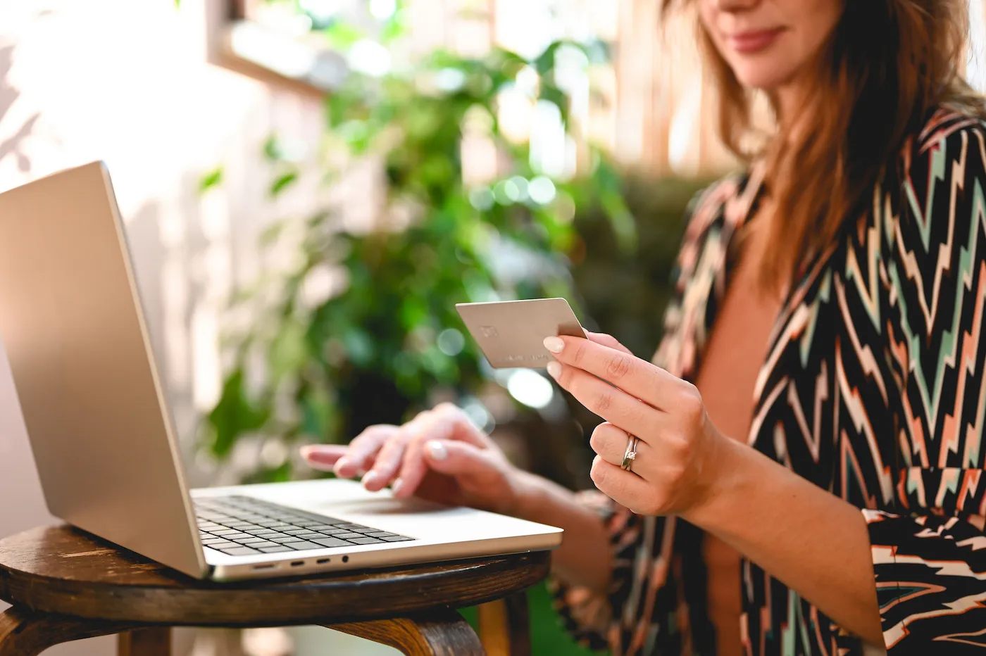 a woman wearing a colorful shirt holds a credit card while typing on her laptop, considering a personal loan or buy now pay later