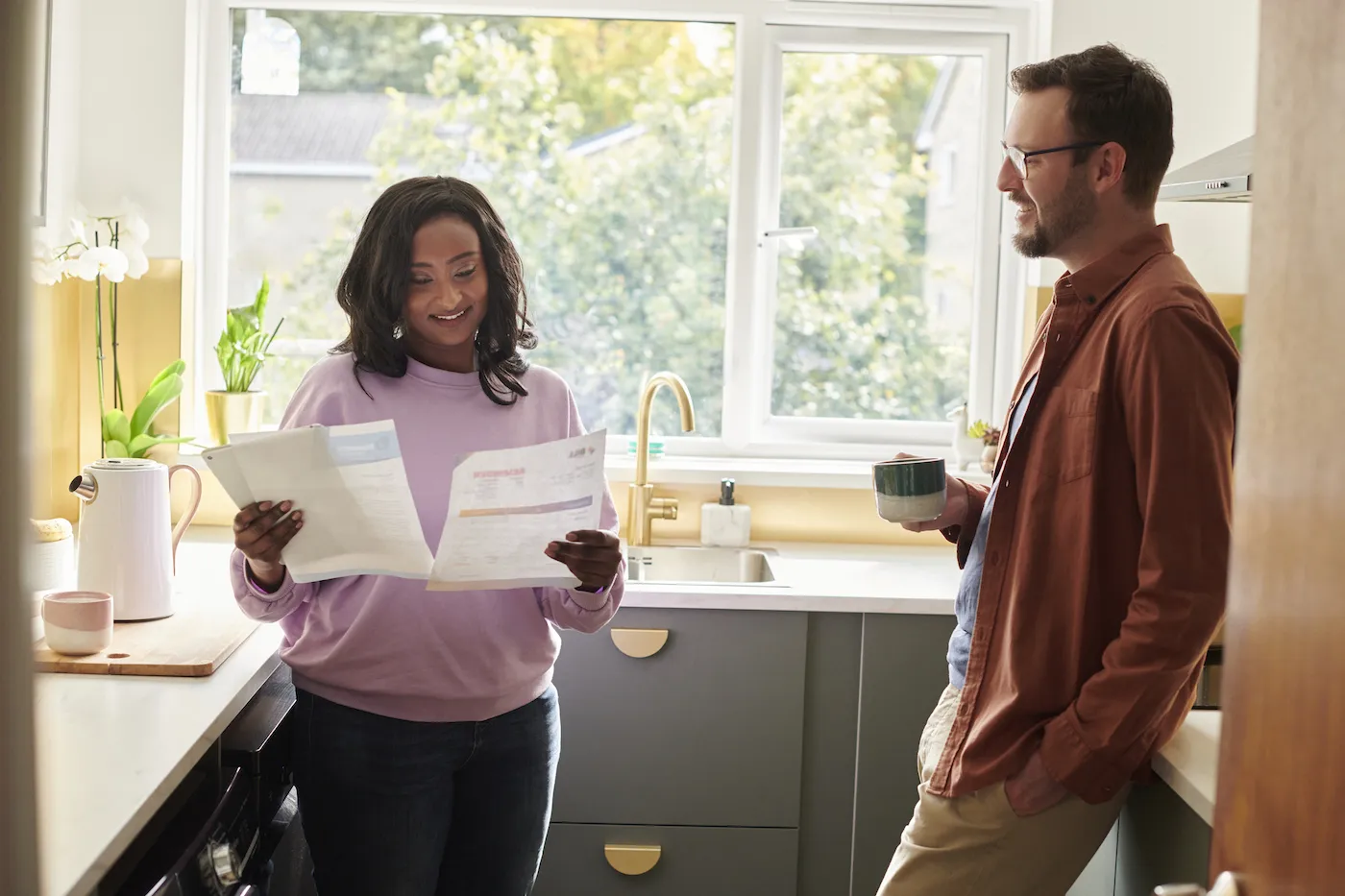 Woman standing in her kitchen with her partner, holding a paper loan offer in each hand to compare them side by side.