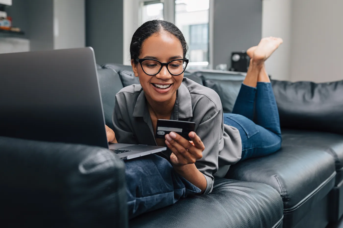 Woman in spectacles lying on a sofa at home holding a credit card.