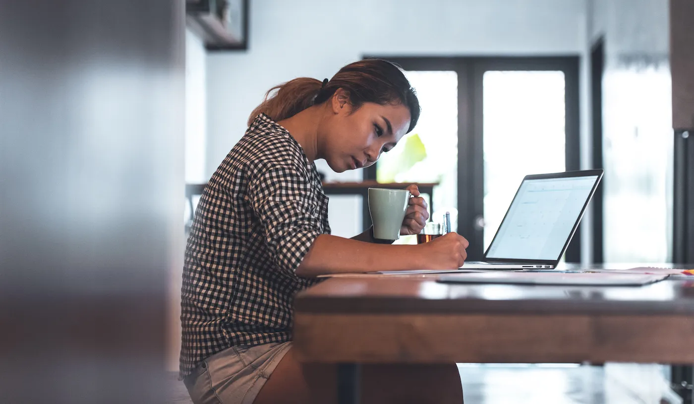 Young woman making monthly budget at table. Female is holding mug while calculating monthly expenses. She is at home.