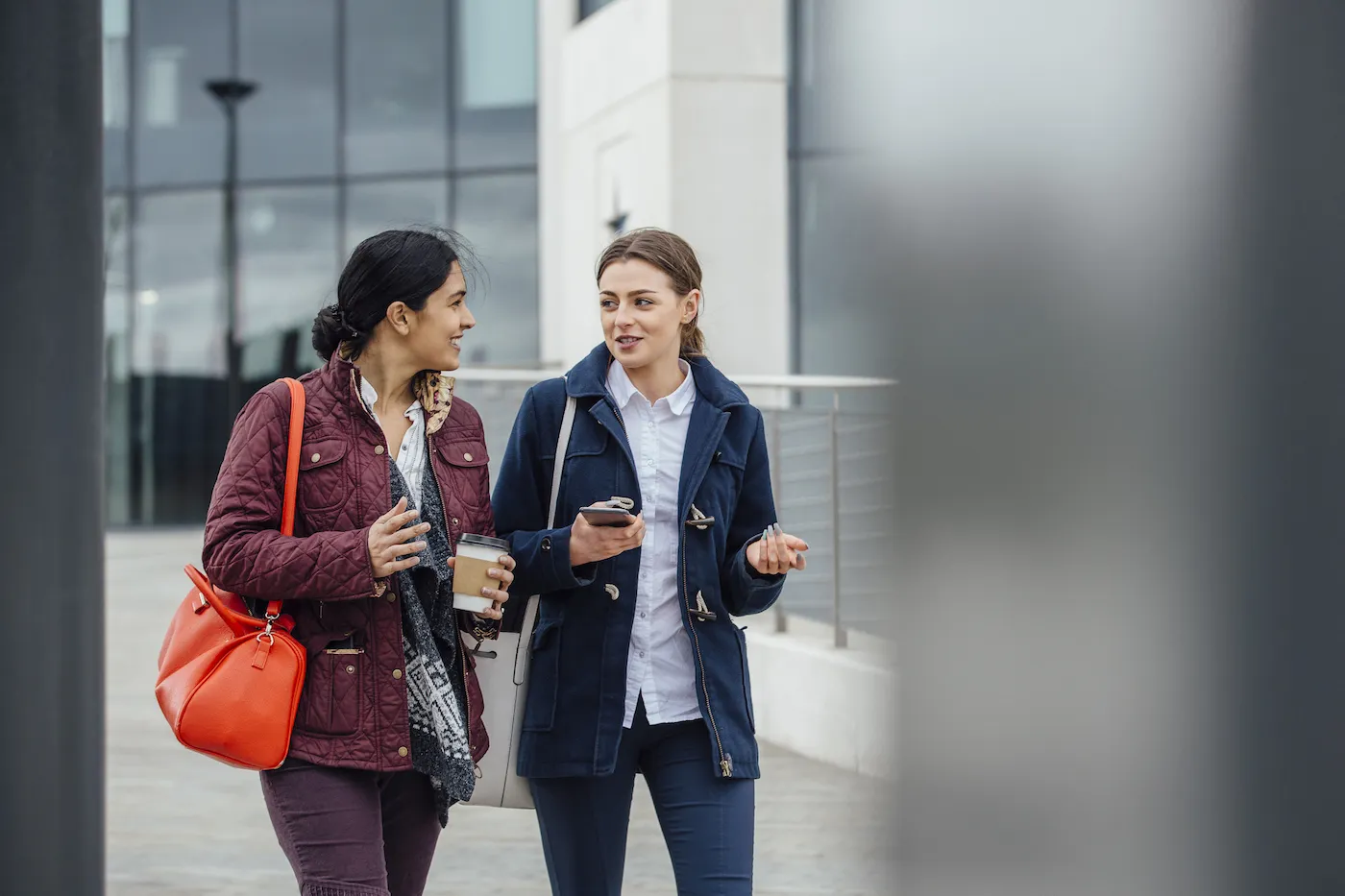 Businesswomen are walking to work together through the city. They are talking and one is holding a coffee while the other is holding a smartphone.