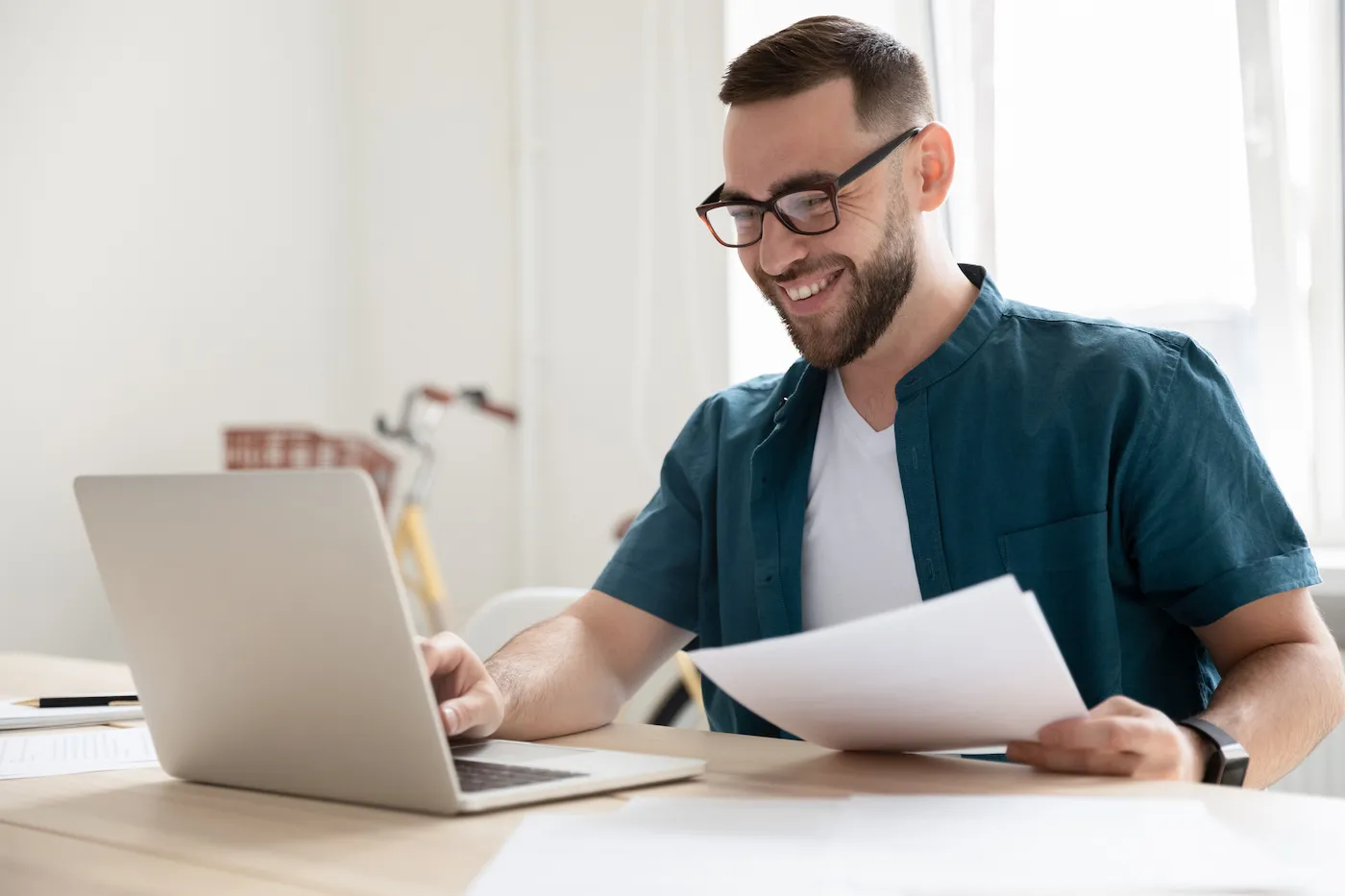 A young man sitting at a desk holding papers and researching HELOCs on his laptop.