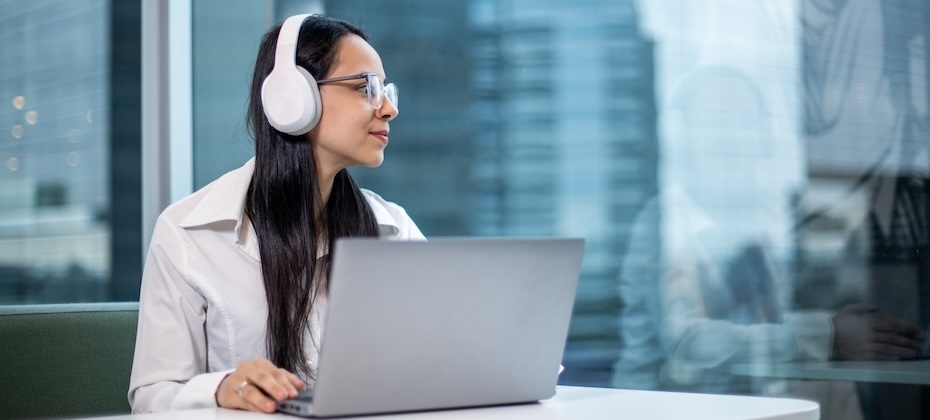 Young woman seated a table with her laptop and headphones, staring out the window where a skyscraper is visible in the background.