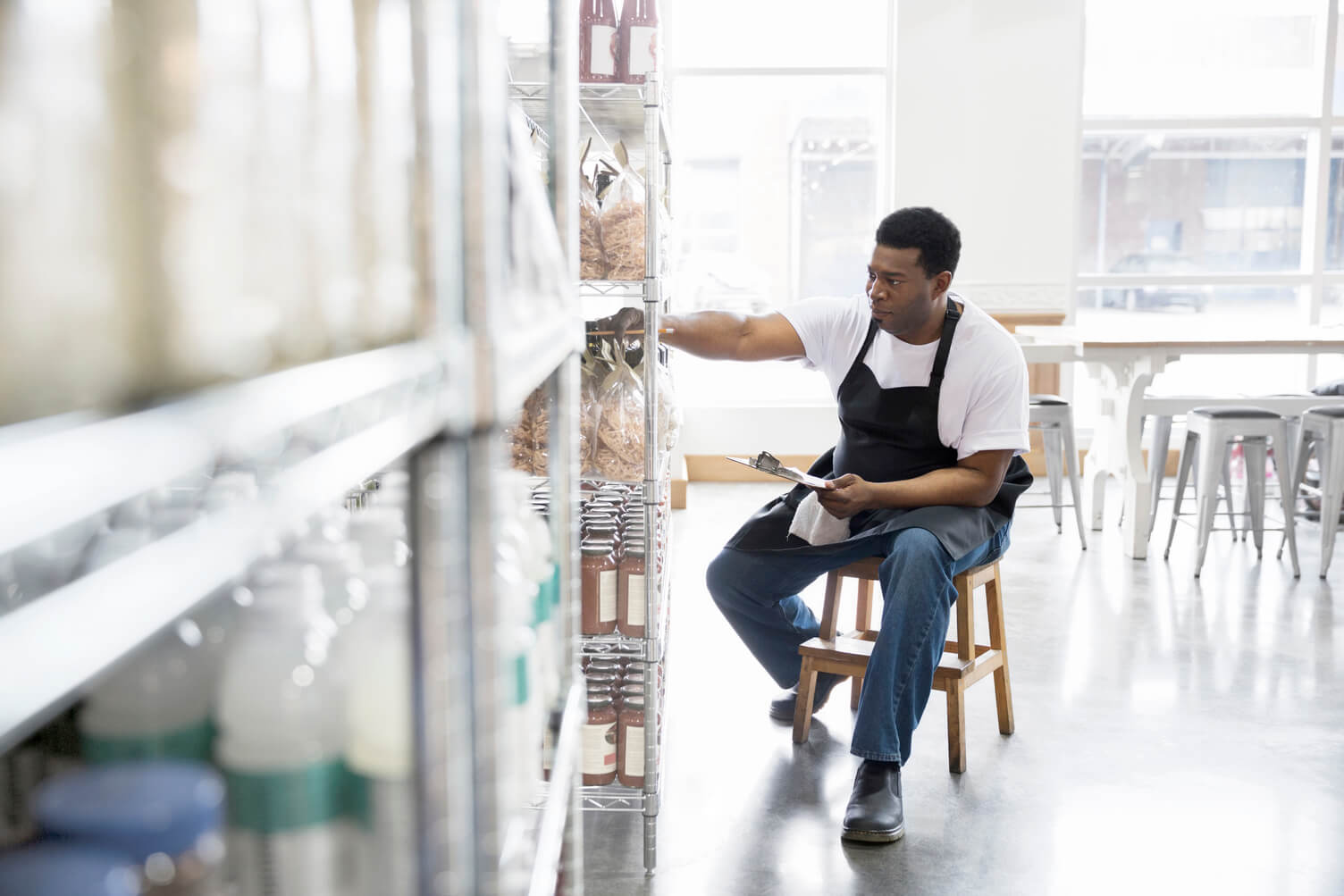 A man sitting on a stool in a bright warehouse, reaching into a shelf