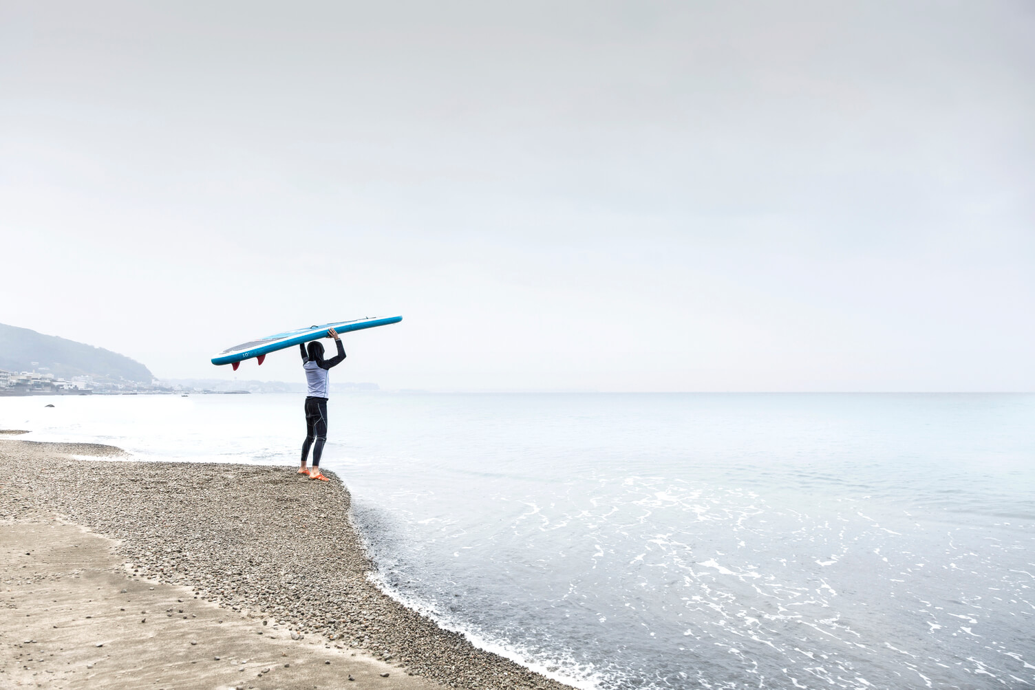 Male surfer standing on shore, holding surfboard over his head, looking out onto water