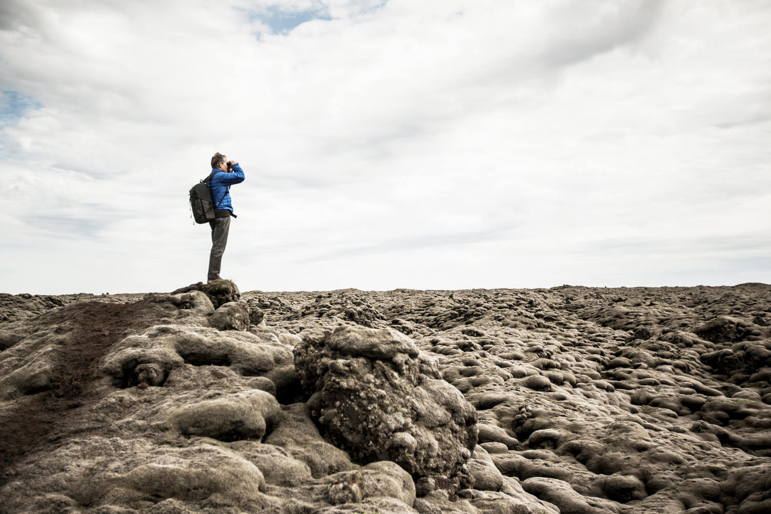 Example image of setting with a male in blue jacket, standing on a boulder, peering through binoculars off into distance