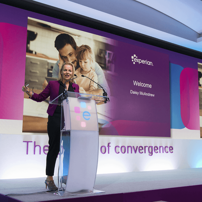 Woman on stage in front of presentation screen, speaking into microphone behind a contemporary, Experian branded lectern